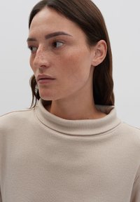 Donna con lunghi capelli castani e lentiggini, che indossa un maglione beige a coste con colletto alto, che guarda a sinistra contro uno sfondo semplice.