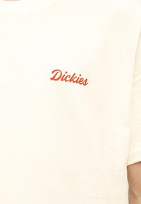 Ύφασμα σε κρεμ χρώμα με μικρό κόκκινο λογότυπο "Dickies" τυπωμένο στην περιοχή του στήθους ενός κοντομάνικου πουκάμισου.