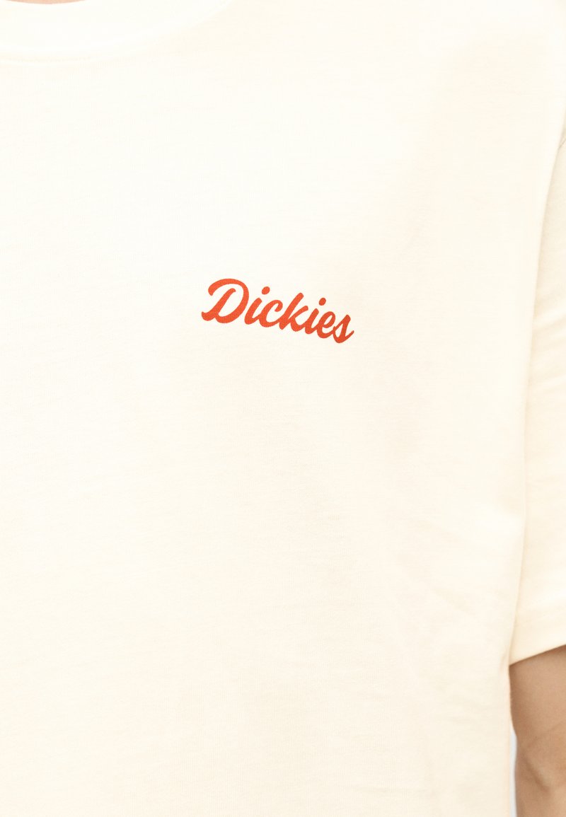 Tela color crema con pequeño logo rojo de "Dickies" impreso en el área del pecho de una camisa de manga corta.