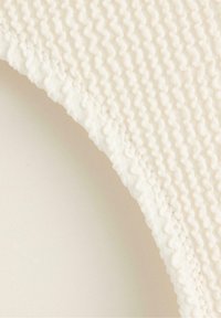 Vit texturerad tyg med vågiga mönster, med en slät kant och sydda detaljer, mot en ljus beige bakgrund.