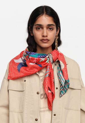 Mujer con cabello largo y oscuro que lleva una chaqueta beige y una bufanda grande de patrón rojo atada alrededor del cuello, con pendientes dorados colgantes.