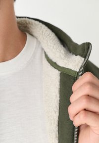 Groene hoodie met rits en een witte, pluche voering, gedeeltelijk open, over een wit T-shirt. Close-up van de stoftextuur en details van de rits.