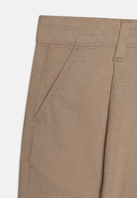 Gros plan sur un pantalon beige avec une poche latérale, une passoire de ceinture et des coutures visibles sur un tissu tissé.