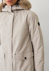 Cappotto invernale beige con cappuccio foderato in pelliccia, dotato di zip frontale e due tasche con bottone a pressione. Tessuto morbido e design minimalista.