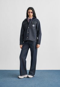 Σκούρο μπλε denim μπουφάν με ορειχάλκινα κουμπιά, λογότυπο στην μπροστινή τσέπη, σετ με τζιν παντελόνια φαρδιάς γραμμής που ταιριάζουν. Μοντέλο φοράει ανοιχτό μοβ πουκάμισο και γυαλιά ηλίου.