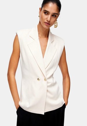 Donna con blazer bianco doppiopetto senza maniche con bottoni dorati e orecchini geometrici dorati, mani nelle tasche dei pantaloni neri, guarda verso destra.