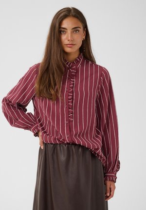 Femme debout, main sur la hanche, portant un chemisier à rayures verticales bordeaux et blanches et une jupe marron foncé, de face.