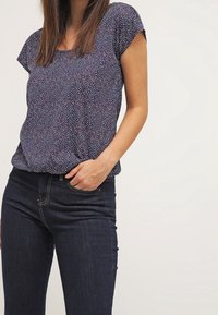 Mujer vistiendo una blusa corta de manga con patrón en azul marino y jeans oscuros, con una mano en el bolsillo frente a un fondo liso.
