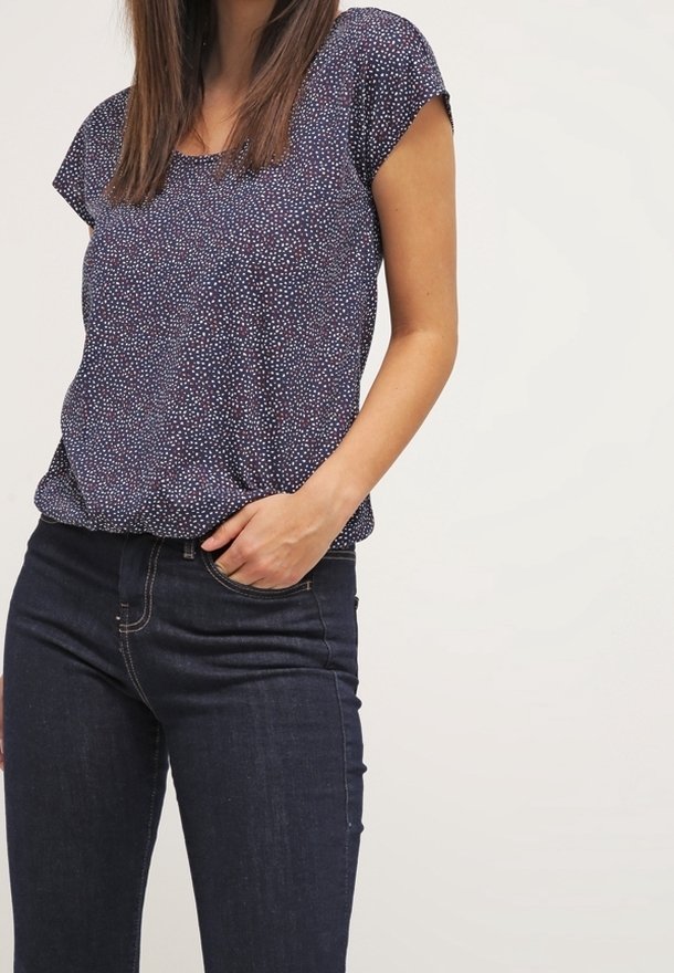 Mujer vistiendo una blusa corta de manga con patrón en azul marino y jeans oscuros, con una mano en el bolsillo frente a un fondo liso.