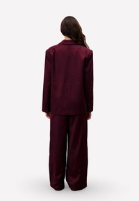 Persoon met lang haar, met de rug naar de camera, gekleed in een oversized donker bordeaux blazer en wijde broek, tegen een effen achtergrond.