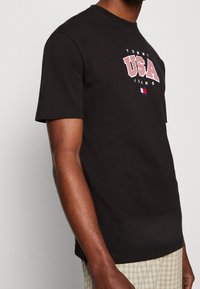 Black cotton short-sleeve t-shirt featuring a white and red graphic "USA" logo with small tricolor flag accent at the hem.