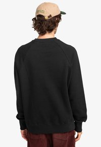 Black sweatshirt with a relaxed fit, ribbed cuffs and hem, featuring a smooth texture. Paired with a tan cap featuring green text.