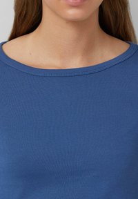 Frau, die ein blaues geripptes Rundhals-Shirt trägt, und dabei ihren Hals, Schlüsselbein sowie einen Teil ihrer Lippen und Haare zeigt.