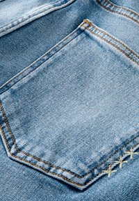Denimtyg i ljusblå färg, med en fyrkantig bakficka med synlig sömnad och en lätt textur. Accent sömnad i ljusgul.