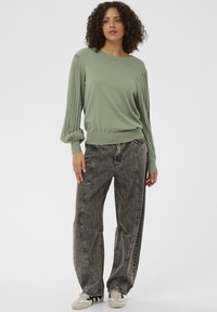 Pulover verde deschis cu mâneci cu dantelă, asortat cu blugi negri largi, cu textură de denim estompat și detalii verticale de cusături.