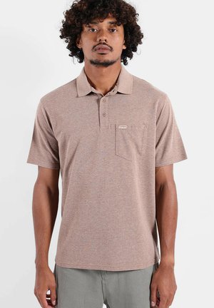 Homme aux cheveux bouclés portant un polo marron à manches courtes avec une poche avant et un pantalon gris clair, debout devant un fond uni.