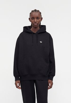 Ung person med flettet hår iført en sort hættetrøje med lille hvidt Adidas-logo, stående mod en ensfarvet lys baggrund.