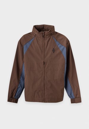 Un mannequin porte une veste marron zippée avec des touches de bleu, une casquette noire, un pantalon noir ample et des baskets épaisses sur un fond uni.