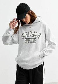 Gray hoodie with "MELROSE AVENUE" printed in black, featuring a front pocket and drawstring hood. Paired with black pants and a black cap.