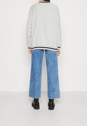 Personne portant un sweat-shirt gris clair avec bordure noire, un jean bleu ample et des chaussures à plateforme noires, debout devant un fond blanc uni.