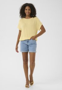 Blouse jaune à manches courtes fabriquée en tissu léger, associée à un short en jean bleu clair retroussé au niveau de l'ourlet. Porté avec des sandales à talons jaunes.