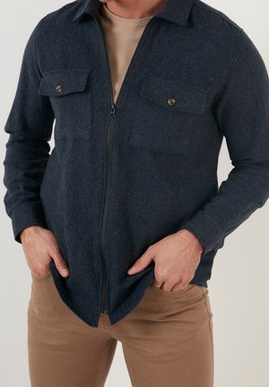 Mann trägt dunkelblaue Wolljacke mit Reißverschluss und Brusttaschen sowie beige Hose, Hände in den Taschen vor hellem Hintergrund.