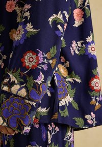 Mornarsko cvjetni materijal s složenim uzorcima u crvenoj, ljubičastoj i zelenoj boji. Ima široke rukave i glatku teksturu, naglašavajući detaljne cvjetne uzorke.