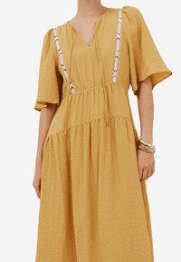 Gelbes, knielanges Kleid aus strukturiertem Stoff, mit V-Ausschnitt, kurzen Ärmeln und weißen Spitzenakzenten in Blumenform an den Trägern.
