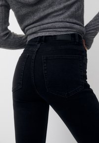 Jeans de mezclilla negra con un ajuste slim. Cuenta con dos bolsillos traseros y una cintura alta. El material tiene un acabado ligeramente texturizado.