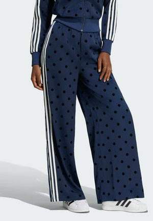 Persona con pantalones de pierna ancha azul marino y chaqueta con lunares negros y rayas laterales blancas, combinados con zapatillas blancas con rayas negras.