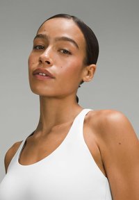 Camiseta blanca sin mangas con escote redondo y tirantes finos, confeccionada con una tela suave y elástica y un diseño ajustado, mostrando un tono de piel natural.