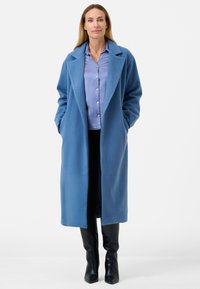 Lungo cappotto blu realizzato in tessuto texture con ampio risvolto, indossato sopra una camicia azzurra chiara. Il modello ha le mani nelle tasche del cappotto. Stivali neri completano il look.