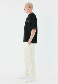 Camiseta negra de manga corta con logo blanco, combinada con pantalones color crema y zapatillas blancas, mostrada de lado sobre un fondo blanco.