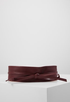 Cinturón ancho de cuero marrón oscuro con una correa delgada atada en un lazo, exhibido sobre una superficie blanca contra un fondo gris liso.