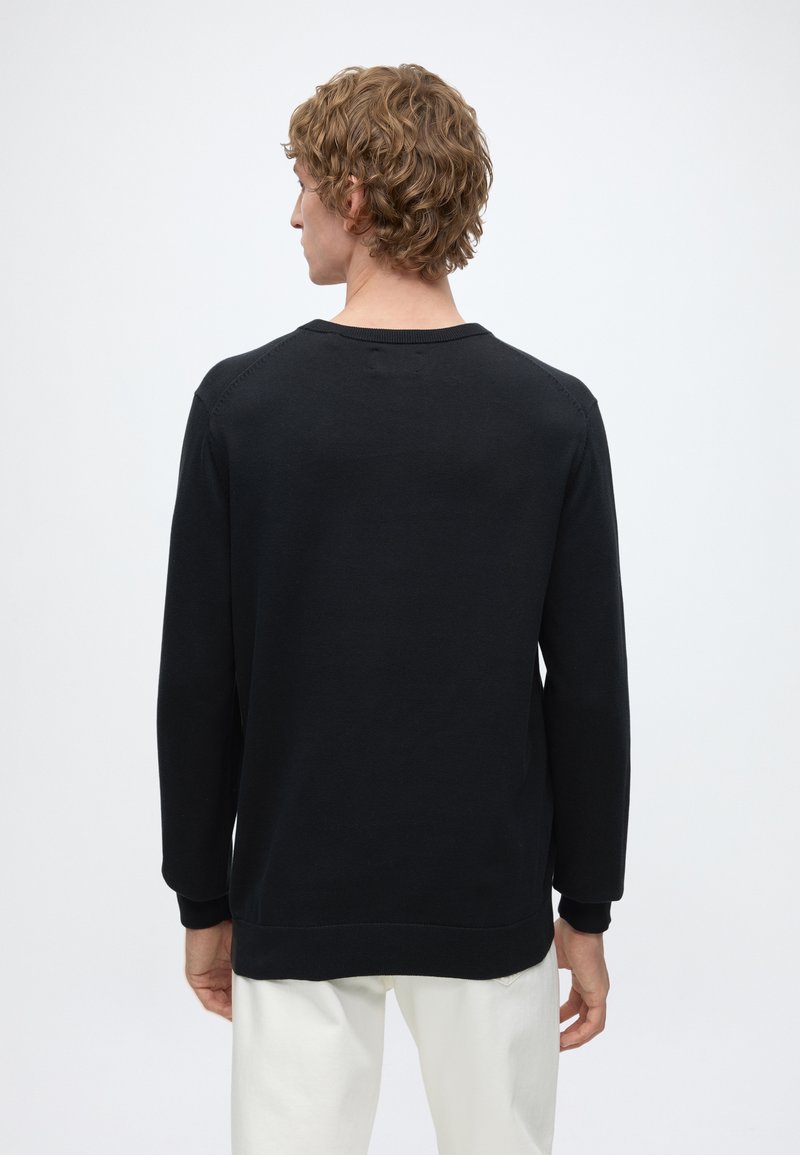 Blond person standing facing away wearing a black long-sleeve shirt and white pants against a plain light background.