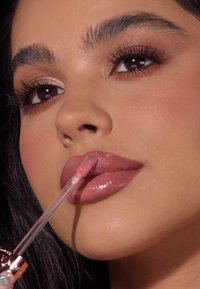 Lipgloss-Applikator in Gebrauch, in einem klaren Röhrchen mit einem verzierten Deckel. Der Gloss hat einen sanften Rosaton und betont glatte, glänzende Lippen.