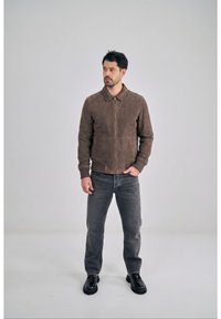 Mężczyzna o ciemnych włosach stojący, ubrany w brązową kurtkę na zamek, wyblakłe czarne dżinsy i czarne buty, na jasnym, jednolitym tle.