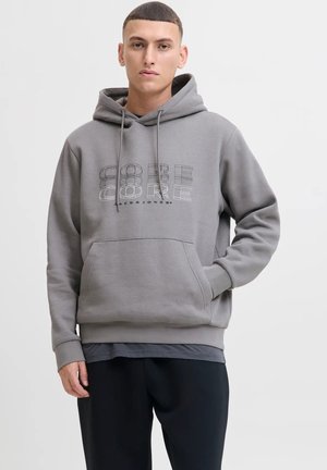Giovane uomo che indossa una felpa grigia con cappuccio e tasca frontale, decorata con un design geometrico del testo "CORE", abbinata a pantaloni neri.