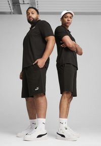 To mænd står ryg mod ryg iført sorte poloer og shorts. De har hvide Puma-sneakers med synlige logodetaljer og rene linjer.