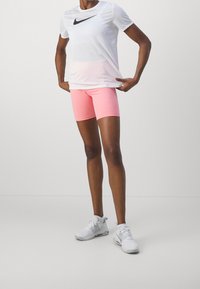 Vit Nike-t-shirt med svart logotyp, matchad med rosa shorts. Modellen bär vita sportskor. Enkla bakgrunder.