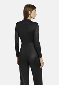 Zwarte jumpsuit met lange mouwen en een getailleerde pasvorm. Het materiaal lijkt soepel en licht glanzend, met een subtiel gestikt patroon.