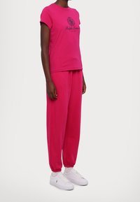 Henkilö, jolla on yllään kirkkaan vaaleanpunainen Ralph Lauren t-paita ja siihen sopivat verkkarit, valkoiset sukat ja valkoiset lenkkarit yksinkertaisella valkoisella taustalla.