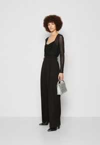 Black sheer long-sleeve top layered over a black tank, paired with high-waisted, wide-leg black trousers. Grey metallic handbag with chain strap.