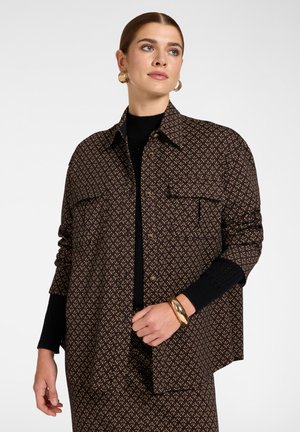Donna che indossa una giacca marrone fantasia con bottoni sopra un maglione nero, con orecchini a cerchio dorati e un braccialetto d’oro, guarda di lato.