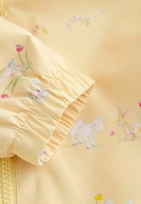 Hele kollane jope pehme kangaga, millel on lille- ja loomamustrid. Elastsed mansetid, nähtav tõmblukk ja sile tekstuur.