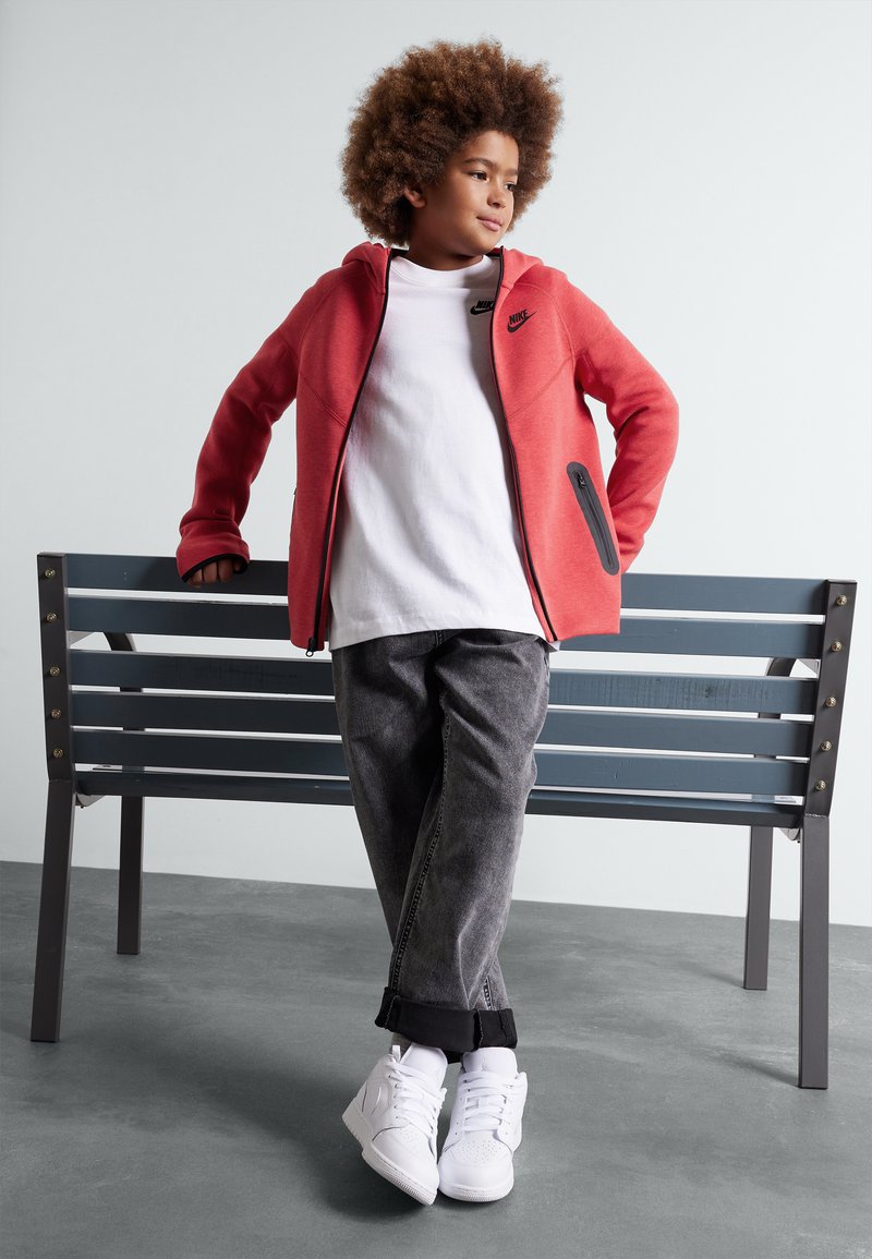 Felpa rossa con zip e dettagli neri, t-shirt bianca, jeans grigi e sneakers bianche. Seduto su una panchina di legno grigia, in una posa rilassata.