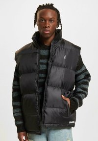 Sort puffer vest med høj krave, lynlås lukning og to sidelommer. Bæres over en sort og grønstribet trøje.