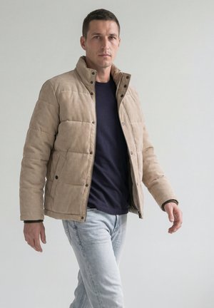 Homme marchant portant une veste matelassée beige par-dessus un t-shirt noir et un jean bleu clair sur un fond gris clair uni.