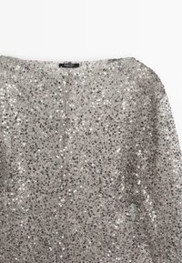 Haut manches longues argenté à sequins avec encolure arrondie, densément couvert de petits sequins circulaires brillants, étiquette visible au col.