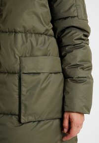 Olivgrüner Puffer-Mantel mit einer glatten, glänzenden Textur. Verfügt über eine aufgesetzte Tasche auf der linken Seite und eine Steppnaht an den Ärmeln.